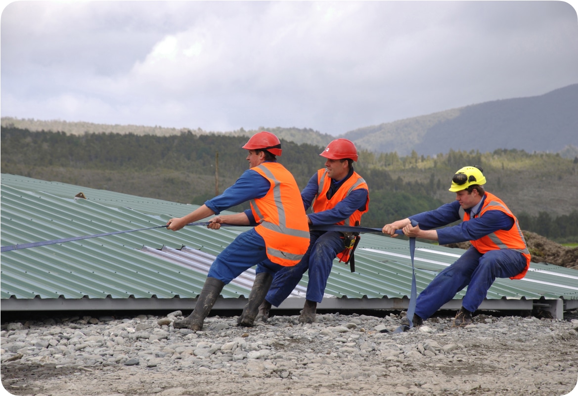 A roofing team pulls a strap to secure metal roofing on a job site.
